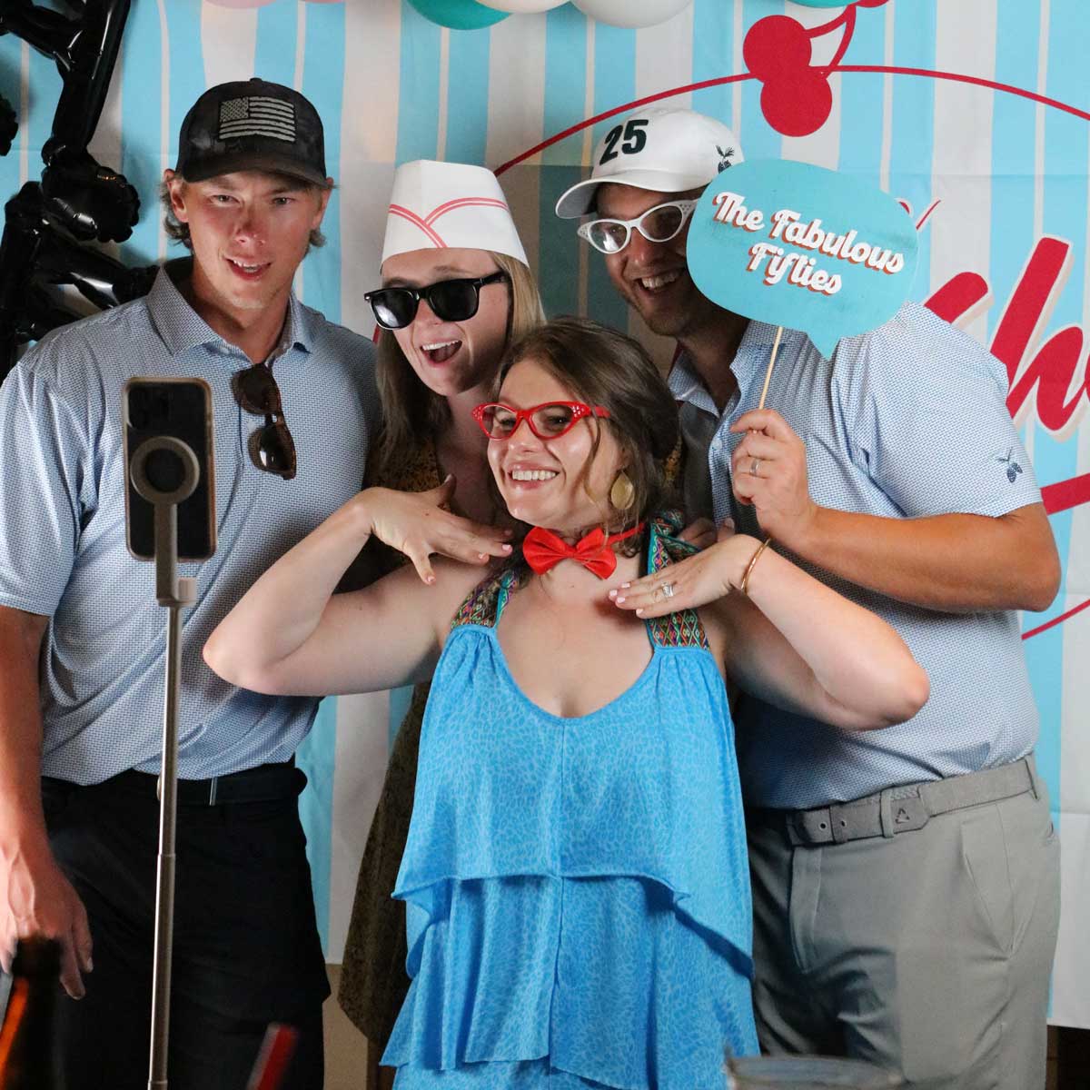 Four people posing for a group selfie in front of a blue and white striped backdrop with red text, one holding a sign saying 'The Fabulous Fifties'.