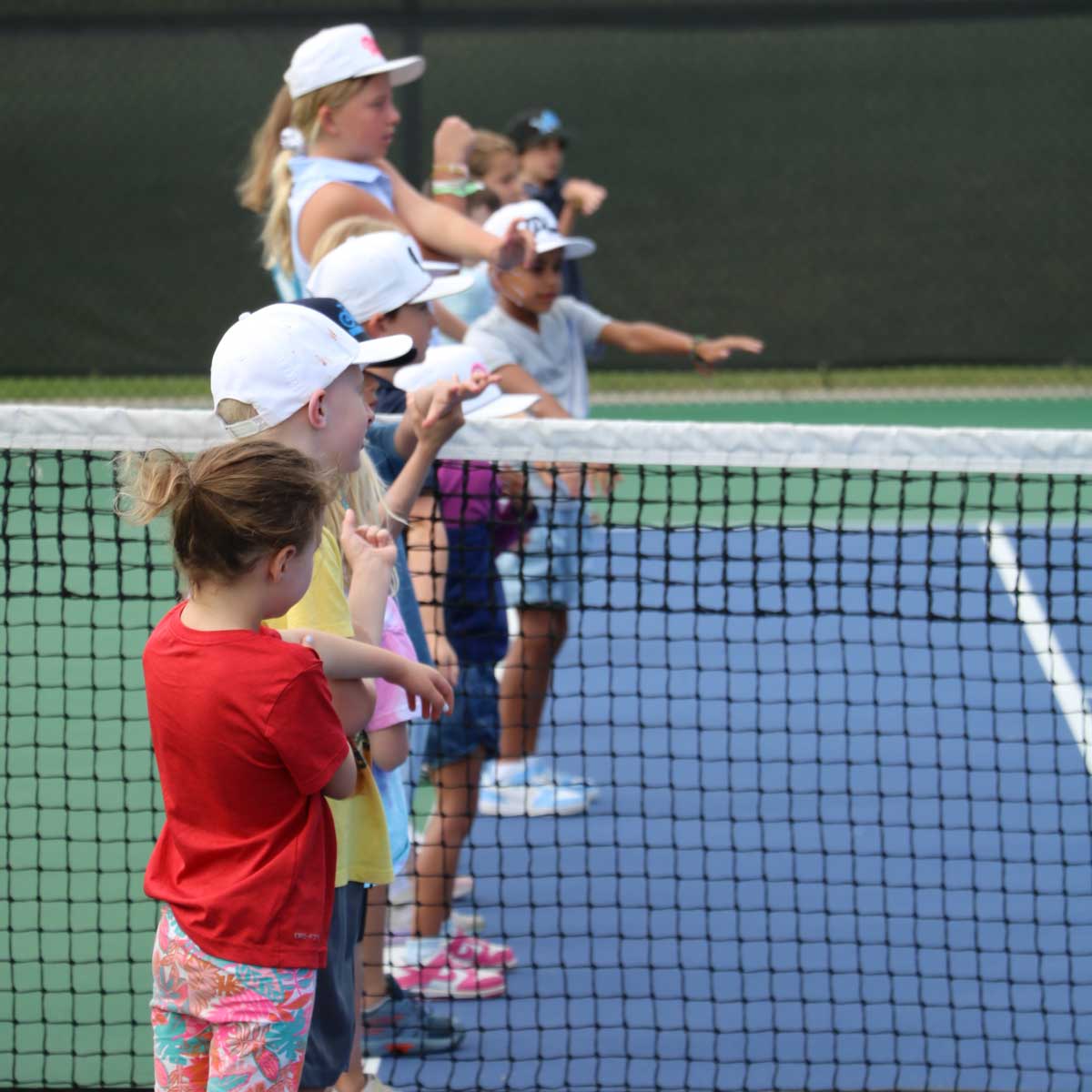 A group of children standing on a tennis court behind the net with arms extended forward.