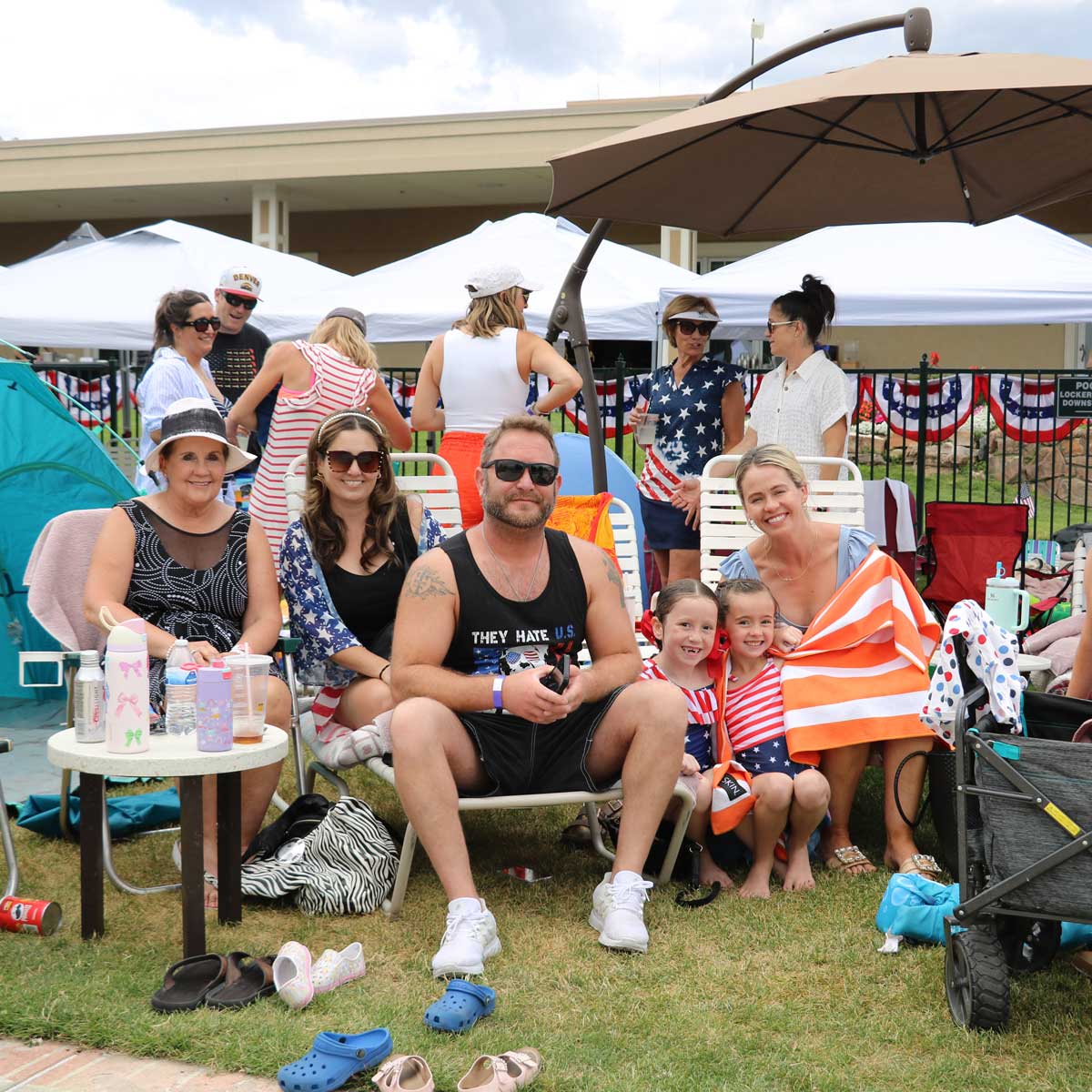 Group of people sitting and standing outdoors under umbrellas and tents, with two young girls in patriotic red, white, and blue outfits, and adults dressed casually, enjoying a social gathering.