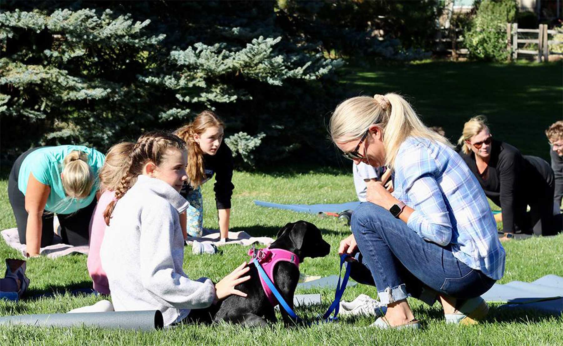 Group of women outdoors on grass with yoga mats and a black dog wearing a pink harness, interacting on a sunny day.