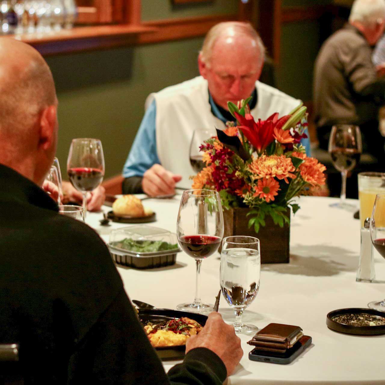 Elderly men dining at a table with wine glasses, a bouquet of flowers in the center, and plates of food.