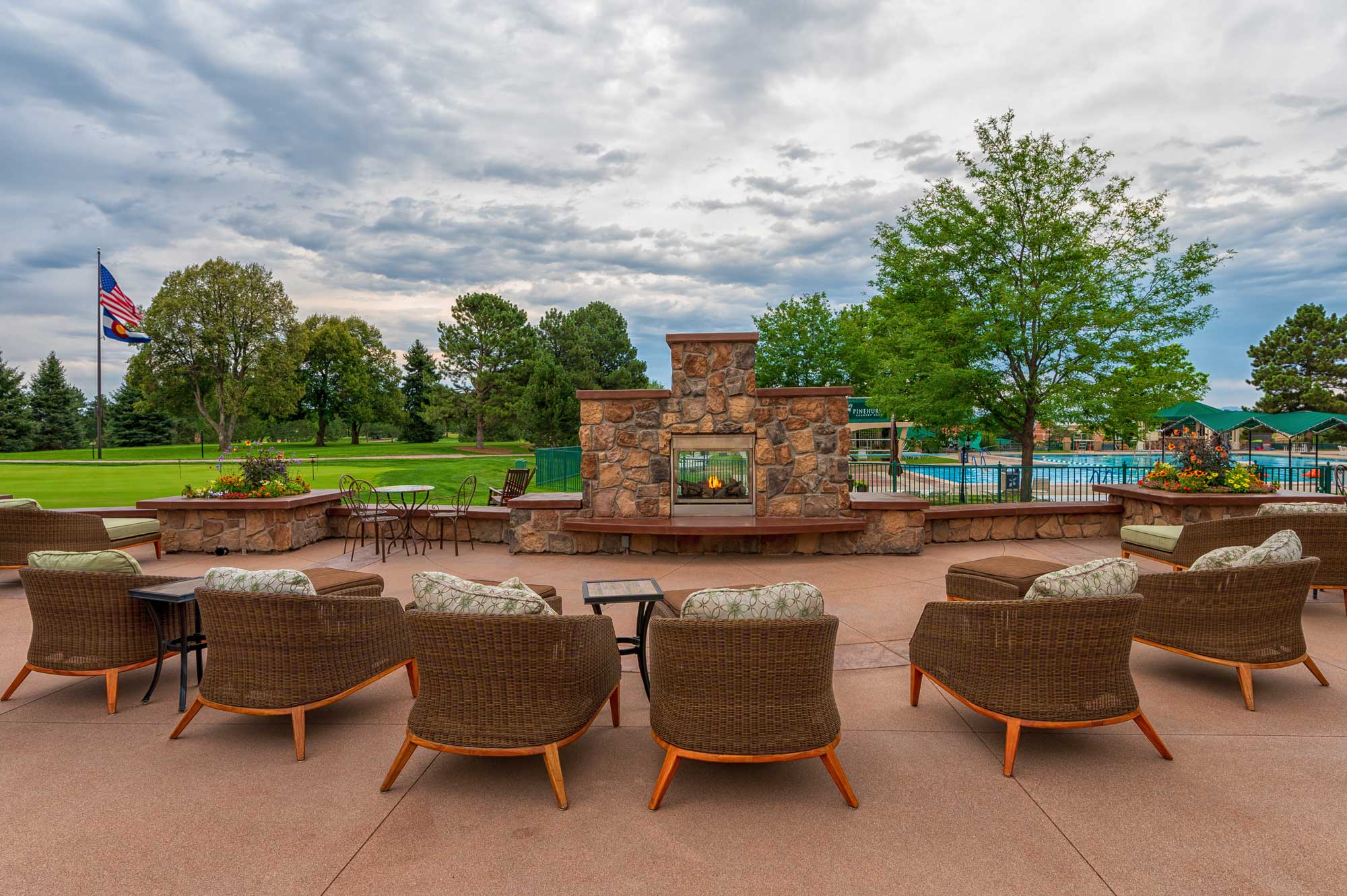 Outdoor patio with wicker chairs arranged in front of a stone fireplace with a fire, overlooking a golf course and swimming pool under a cloudy sky.