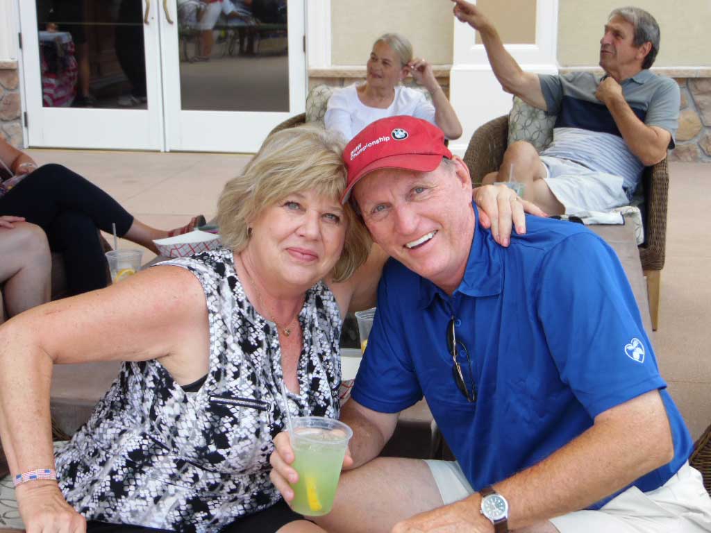 Smiling middle-aged couple sitting closely, woman holding a drink with a lemon slice, man wearing a red cap and blue shirt.