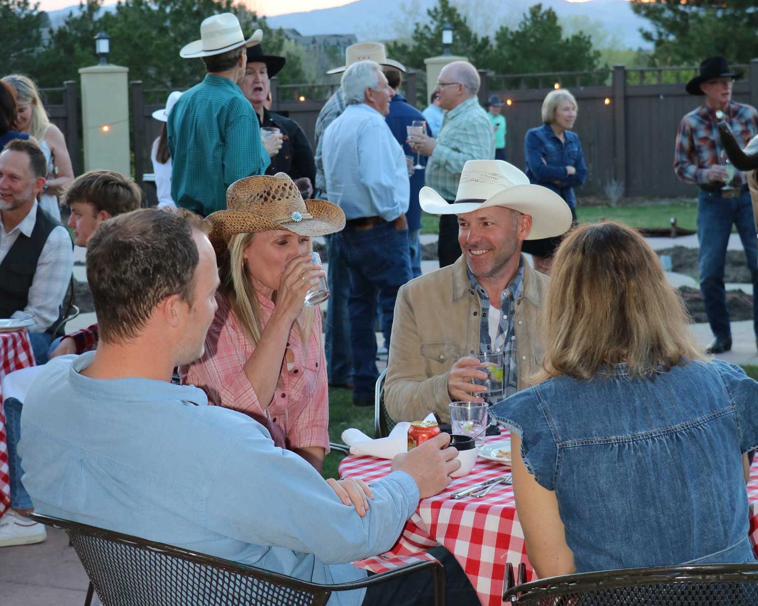 Group of people socializing outdoors at a table with a red and white checkered tablecloth, some wearing cowboy hats and casual clothing.