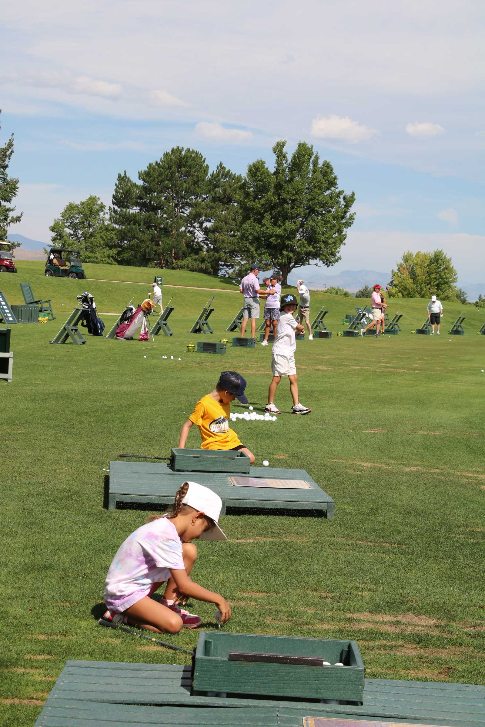 Children practicing golf swings and picking up golf balls on a sunny driving range with trees and adults in the background.