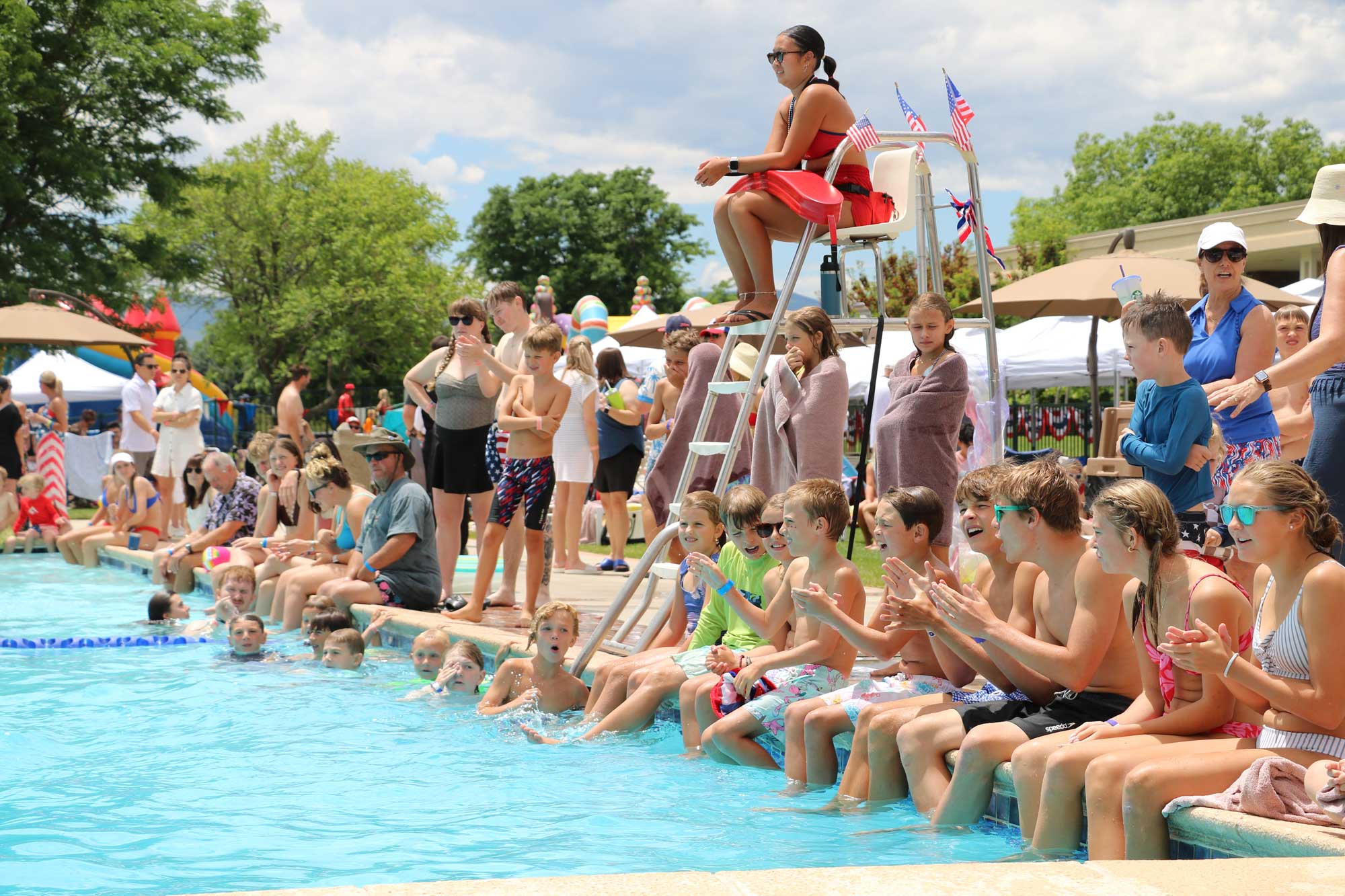 Children sitting and clapping by the edge of an outdoor pool with a lifeguard seated on a chair decorated with small American flags.