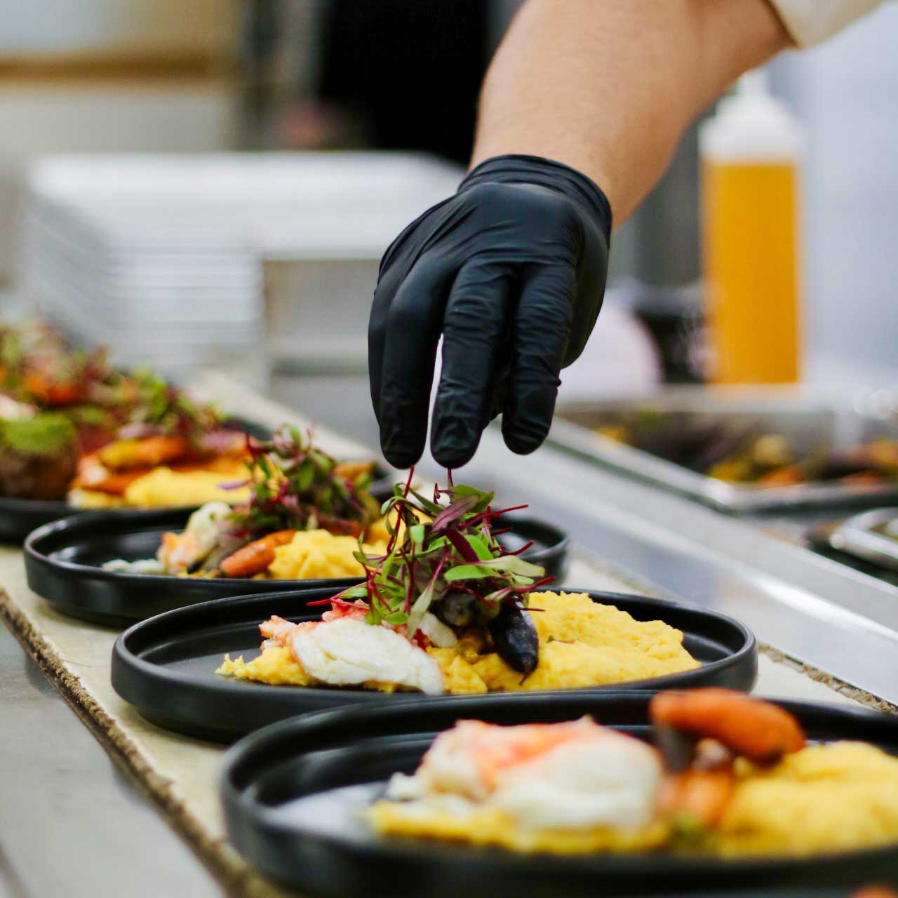 Chef wearing black gloves placing microgreens on plated scrambled eggs with seafood in a professional kitchen.