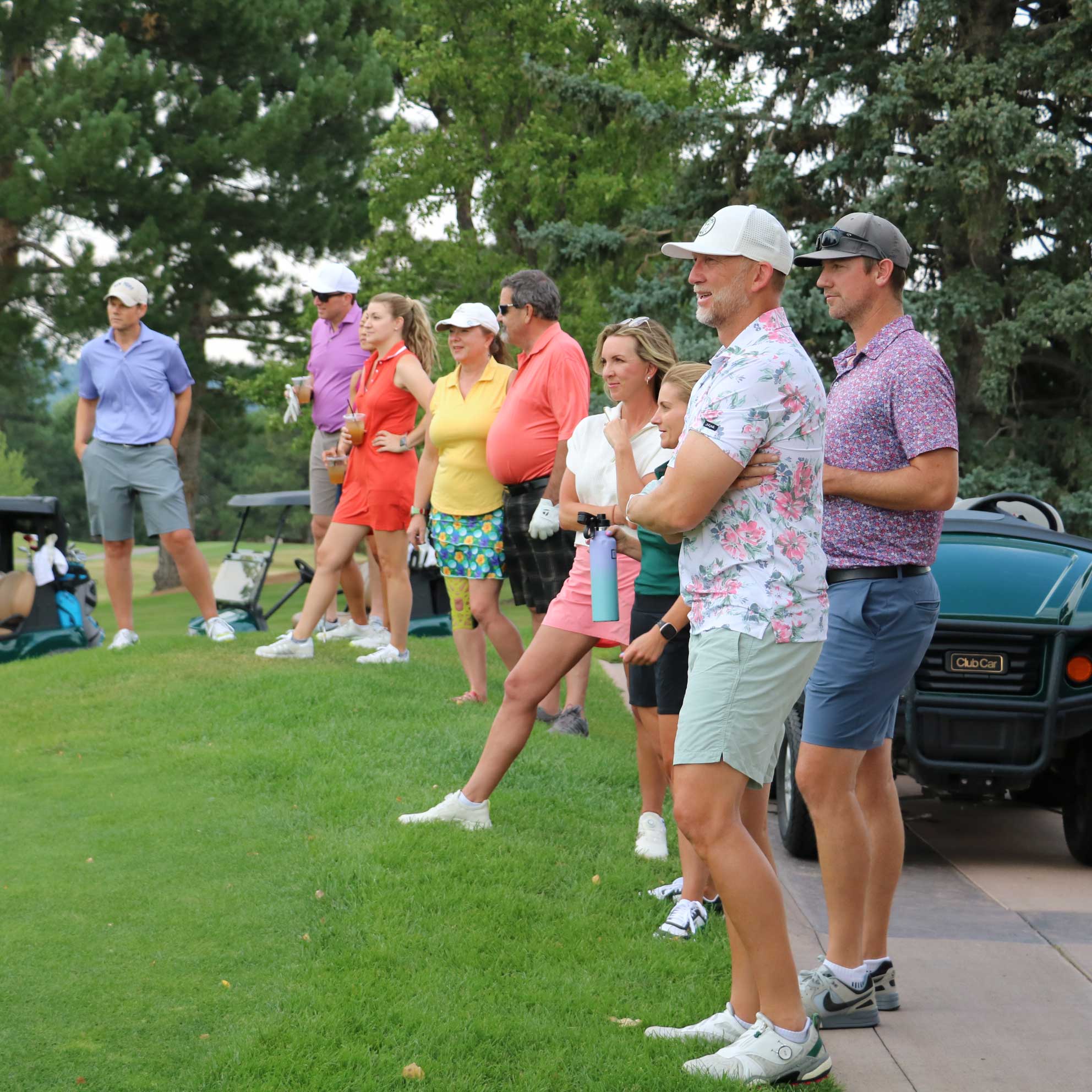 Group of people standing on a golf course near golf carts, watching something off-frame, with trees in the background.