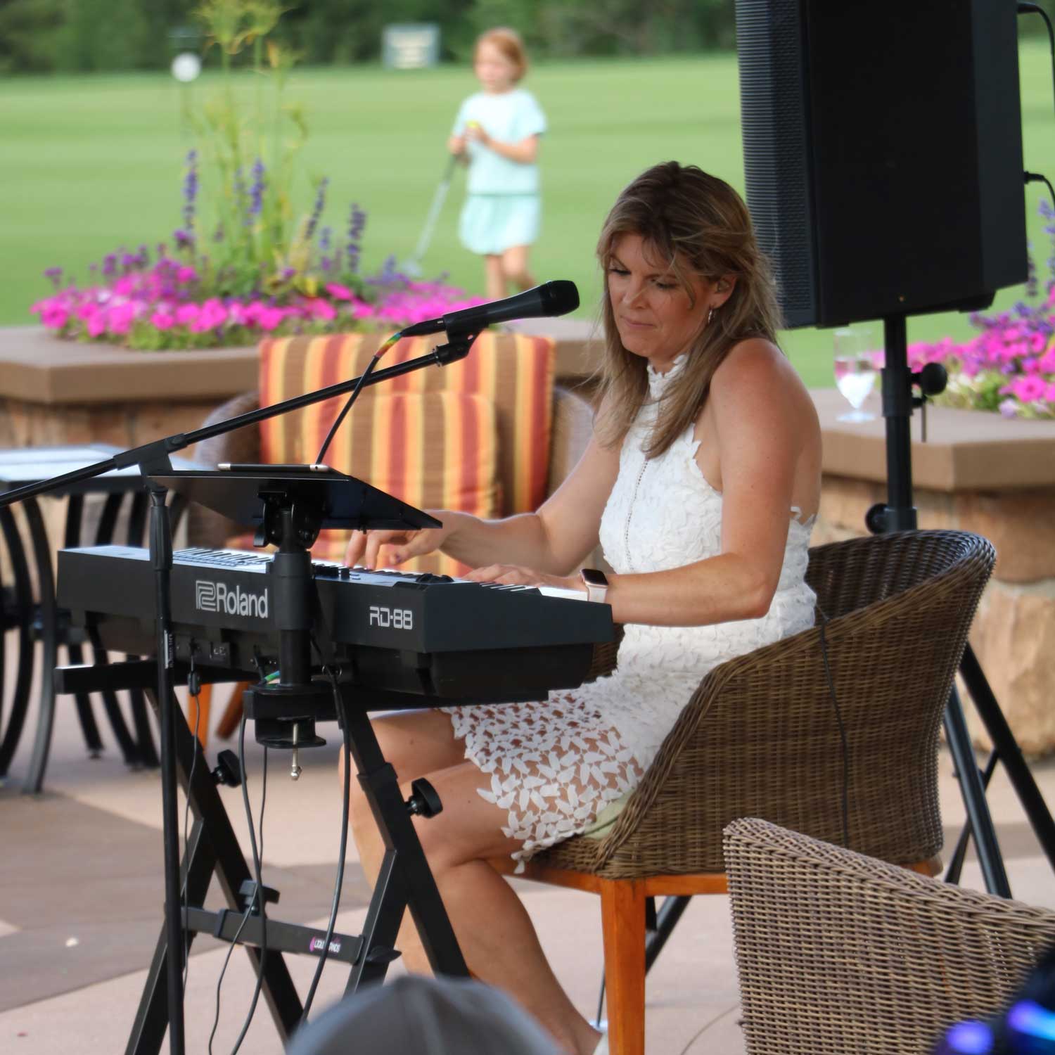 Woman in white sleeveless dress playing a Roland keyboard with a microphone in front, outdoors with flowers and green lawn in the background.