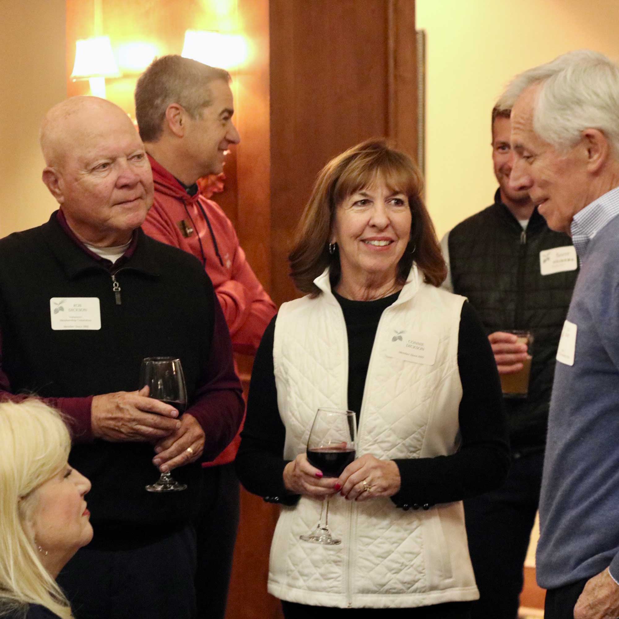 Group of five adults engaged in conversation at an indoor social event, two holding glasses of red wine.