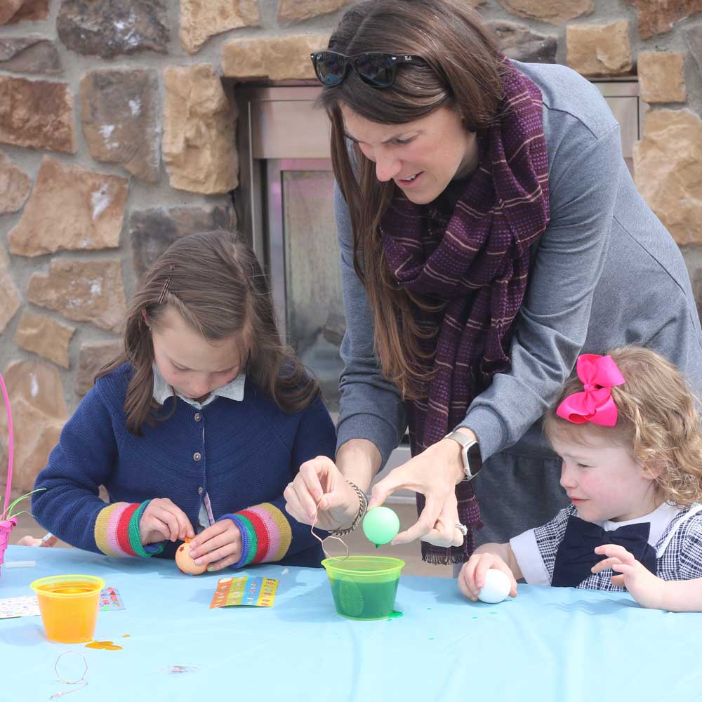 Woman helping two young girls dye Easter eggs at a table covered with a blue cloth.