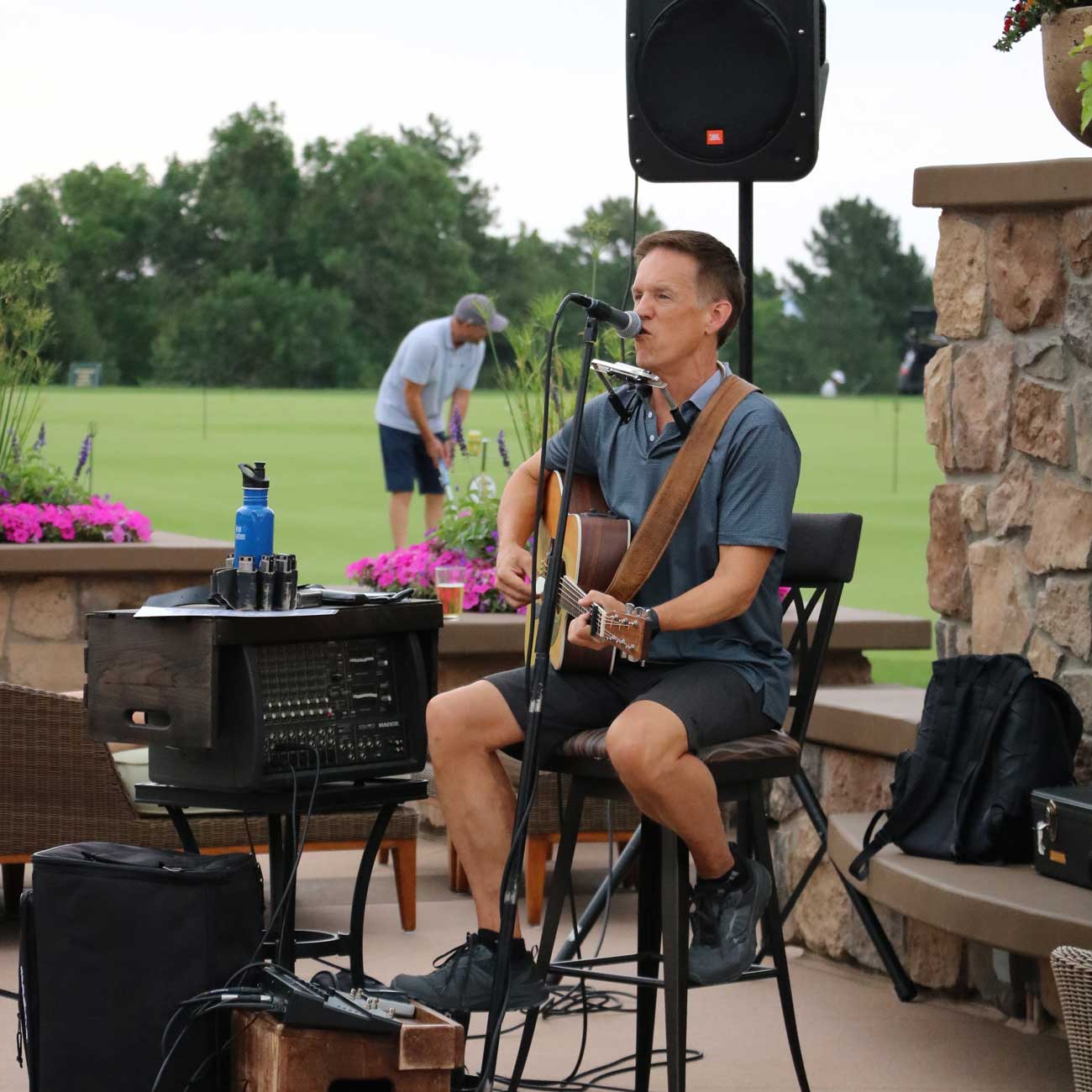 Man playing guitar and singing into a microphone outdoors with sound equipment nearby and a person golfing in the background.