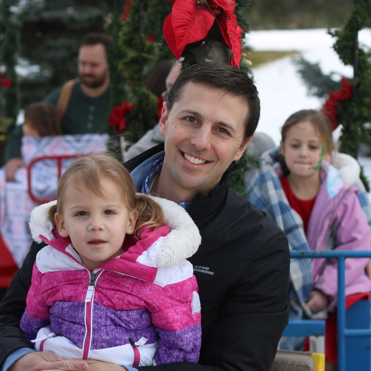 Man smiling and holding a young girl in a pink and purple winter jacket, with other children nearby on a decorated holiday ride outdoors.