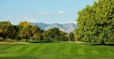 Green golf fairway lined with trees under a blue sky and mountains in the background.