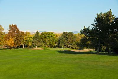 Golf course fairway with green grass, surrounded by trees with some autumn foliage under clear blue sky.