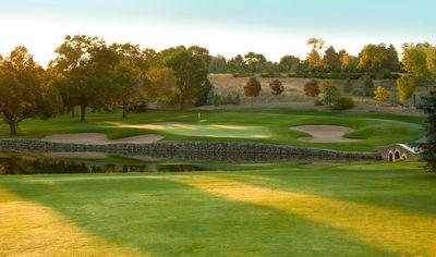Golf course green with sand bunkers, stone wall, and trees under a clear sky.