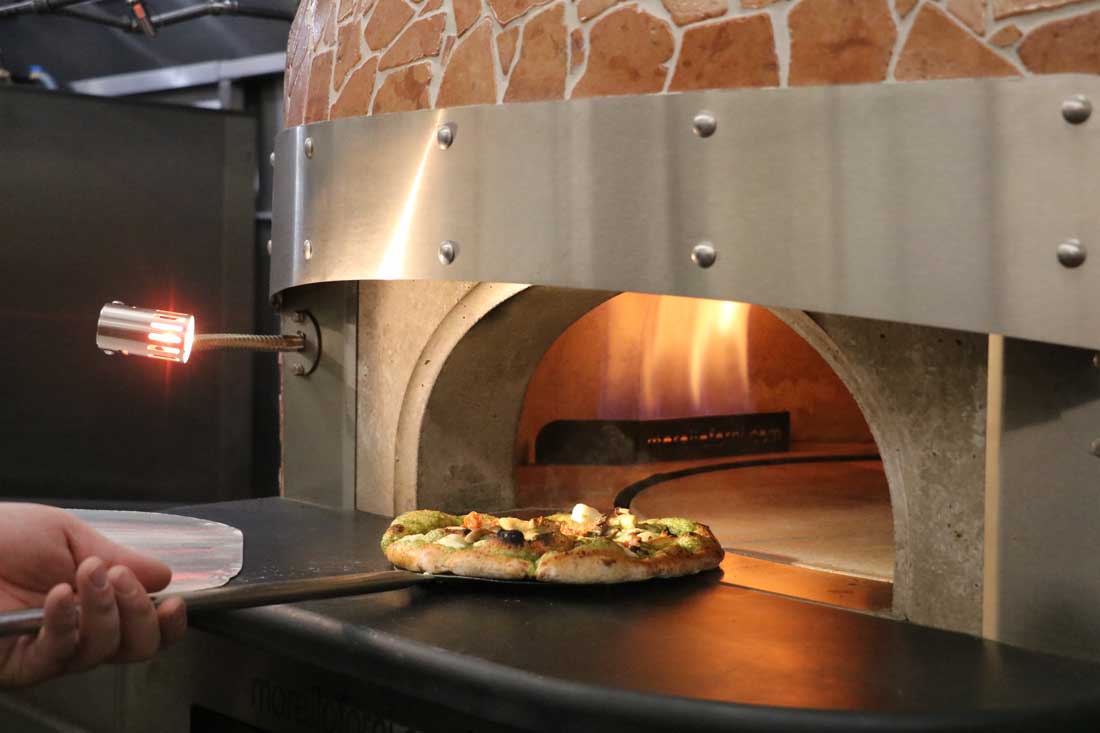 Person placing a pizza with green pesto and cheese inside a brick pizza oven with a visible flame.