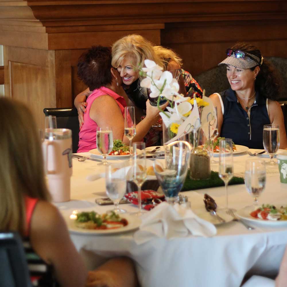 Group of women seated at a dining table with white tablecloth, enjoying a meal and sharing smiles and hugs in a warm, wood-paneled room.
