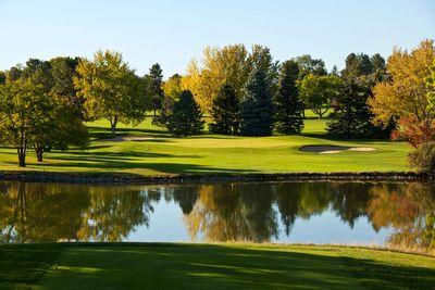 Golf course green bordered by trees and sand bunkers reflected in a calm water hazard under a clear sky.