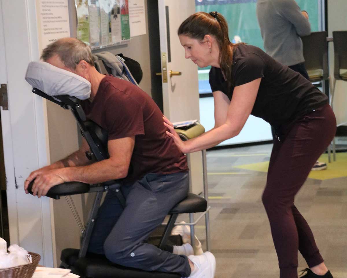 A woman giving a seated man a back massage on a portable massage chair indoors.