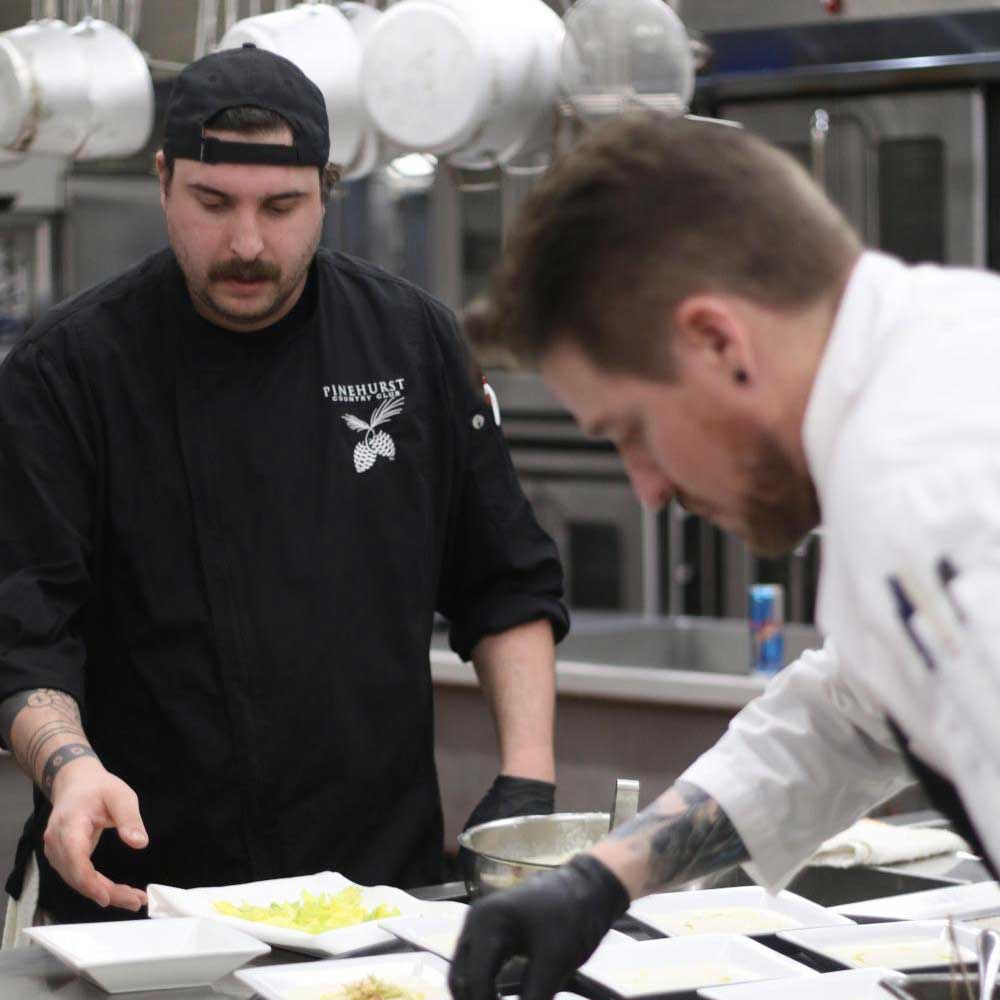 Two chefs preparing plates with food in a professional kitchen, one wearing a Pinehurst Country Club uniform.