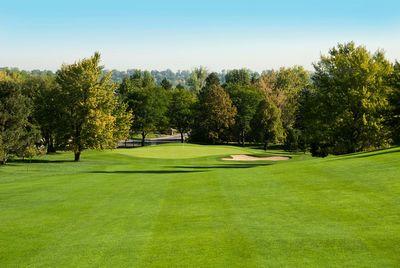 Golf course fairway with sand bunker and green surrounded by trees under a clear blue sky.