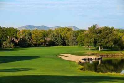Golf course fairway with sand bunker and pond beside it, surrounded by trees and mountains in the background under a partly cloudy sky.