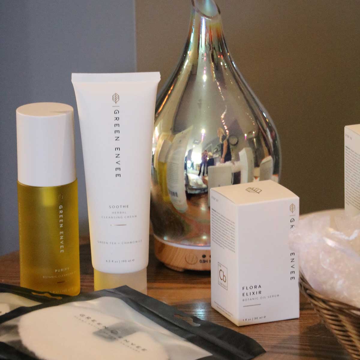 A selection of Green Envee skincare products including Purify botanical plant oil, Soothe herbal cleansing cream, Flora Elixir botanic oil serum, displayed on a wooden surface near a reflective diffuser.
