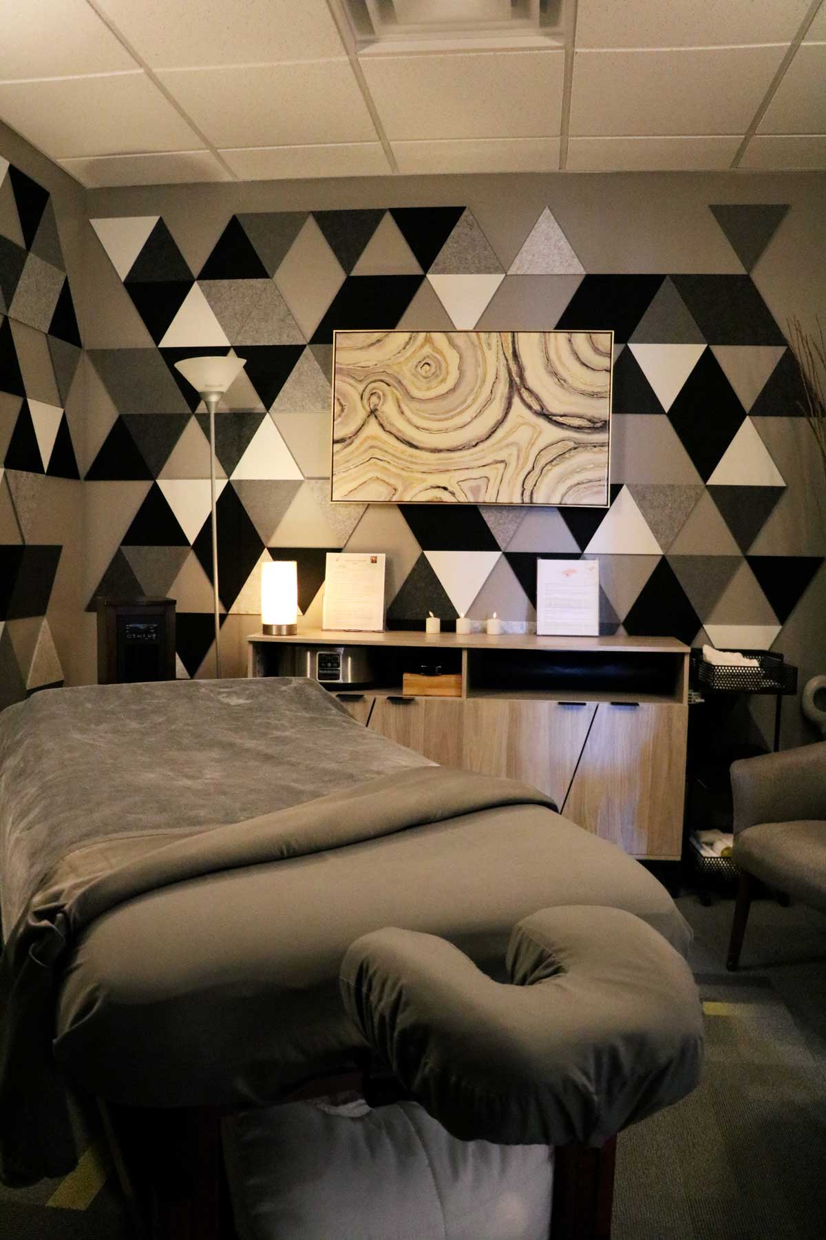 Massage room with a table covered in gray sheets, geometric patterned walls, soft lighting, and a wooden cabinet with candles and documents.