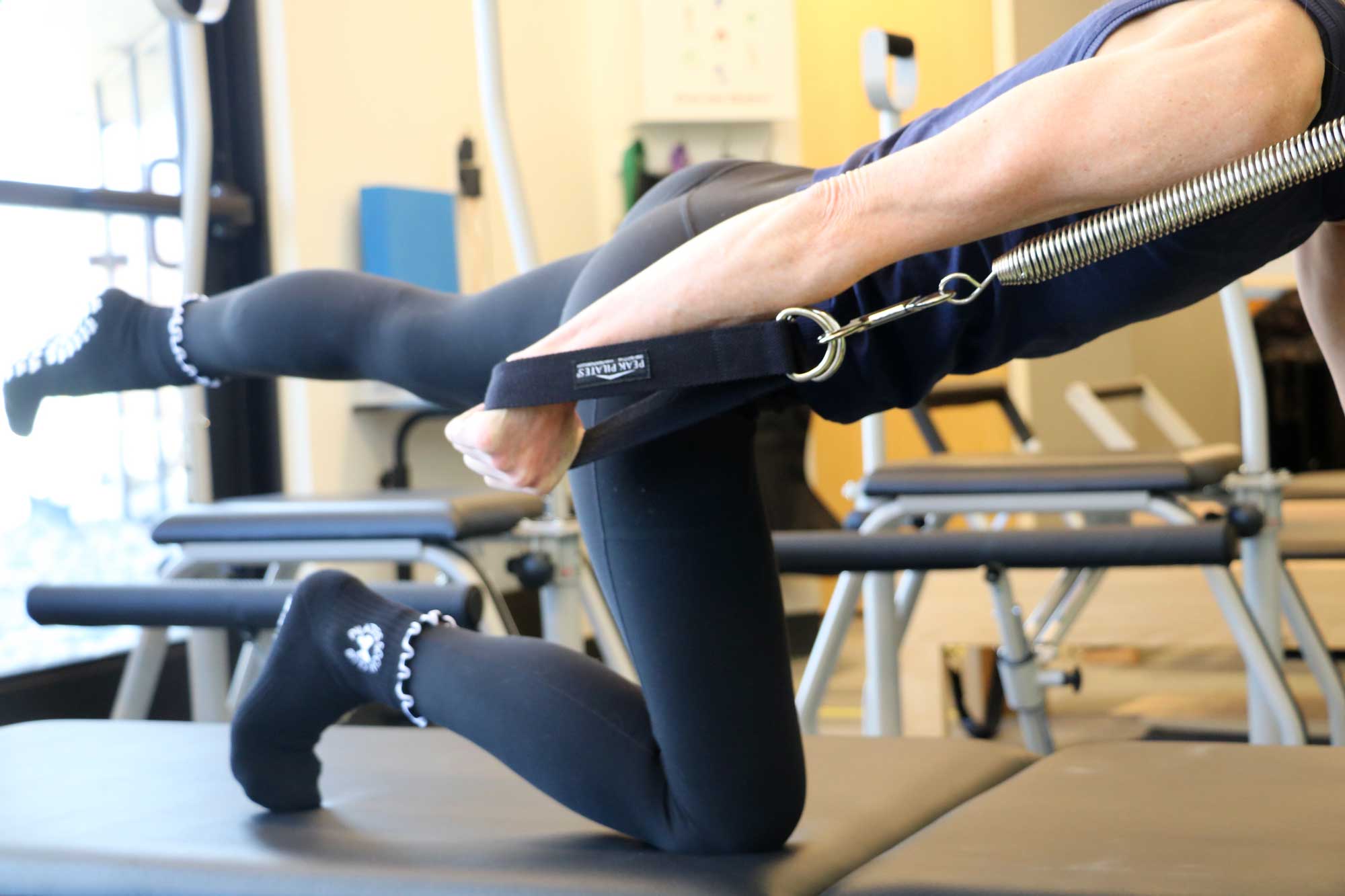 Person performing a Pilates exercise using a spring resistance strap while kneeling on a mat.