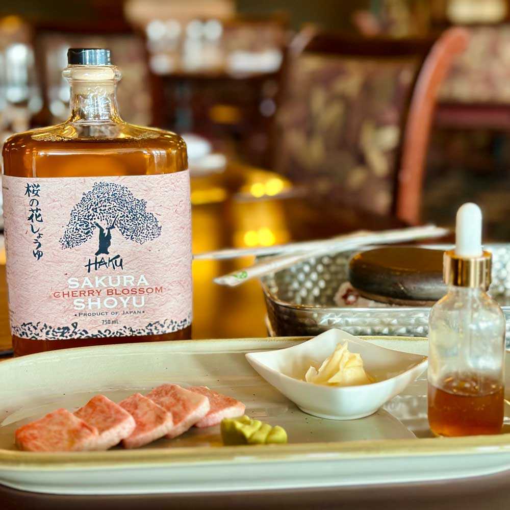 Plate with sliced raw fish, wasabi, and pickled ginger, accompanied by a bottle of Sakura Cherry Blossom Shoyu soy sauce and a small dropper bottle with sauce.