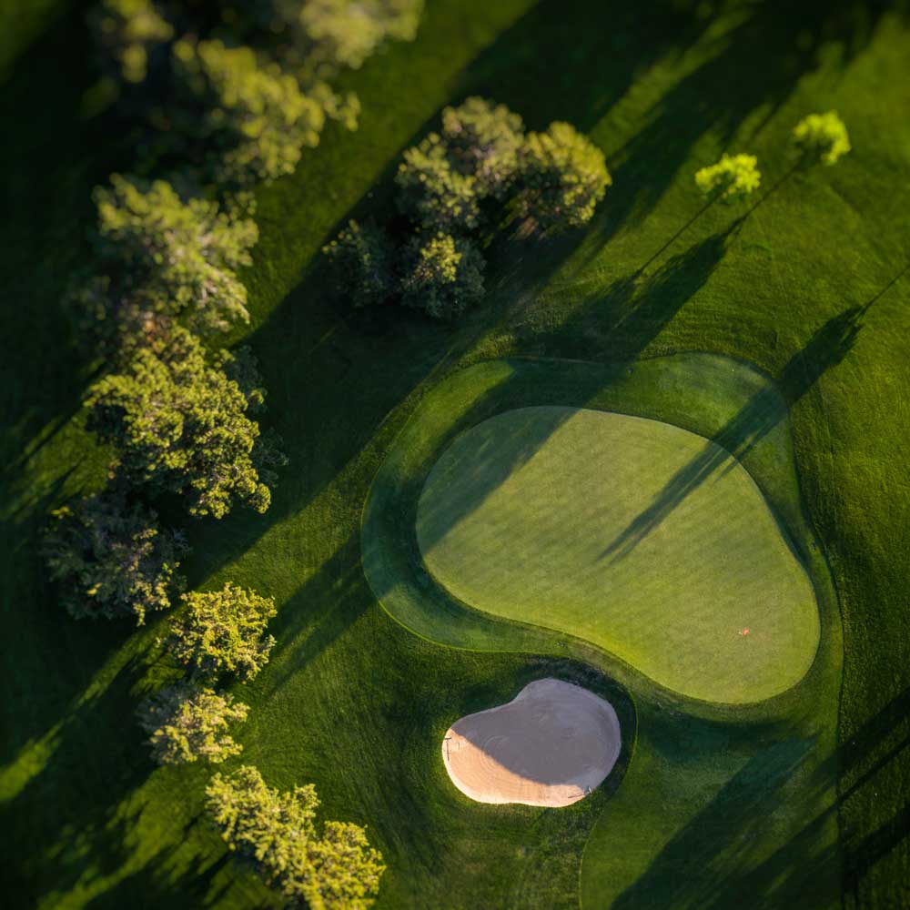 Aerial view of a golf green with a sand bunker and surrounding trees casting long shadows on the grass.