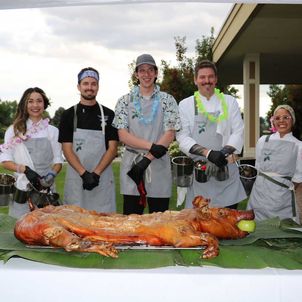 Five people wearing aprons and gloves stand behind a roasted whole pig with an apple in its mouth on a banana leaf-covered table.