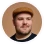 Man with a beard wearing a brown cap and a dark shirt, looking slightly to the side against a white background.