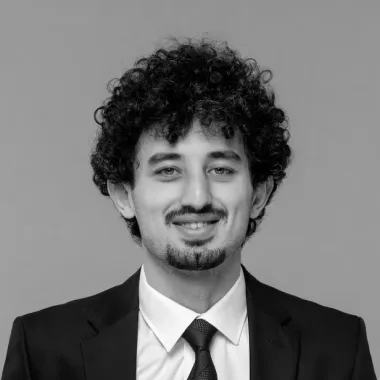 Smiling man with curly hair, beard, and mustache wearing a black suit and tie against a plain background.