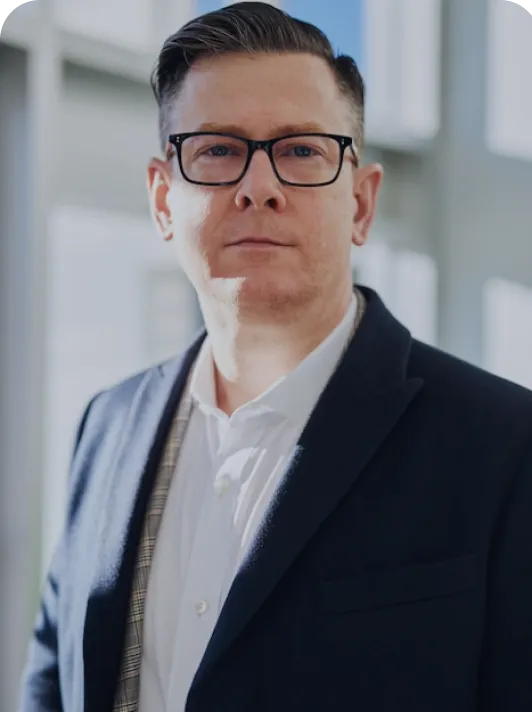 Portrait of a man wearing glasses and a dark blazer over a white shirt, standing indoors with a blurred window background.