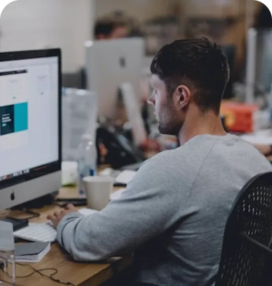 Man in a gray sweater working on a desktop computer in an office setting.