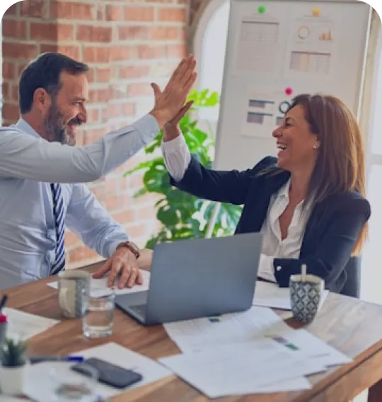 Two business colleagues smiling and giving each other a high five across a desk with a laptop, documents, and coffee mugs.