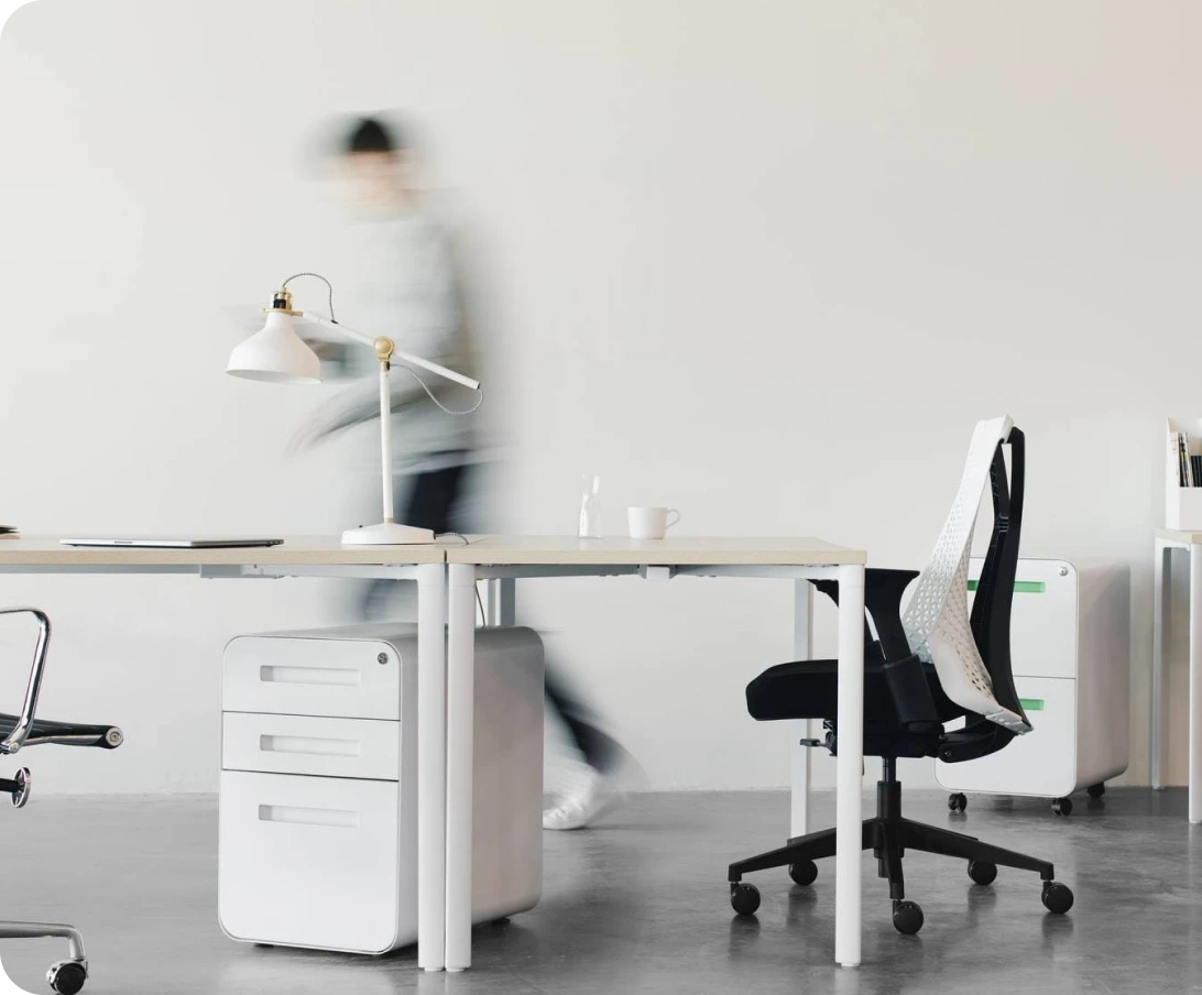 Minimalist office space with white desks, modern chairs, a white desk lamp, and a blurred figure walking in the background.