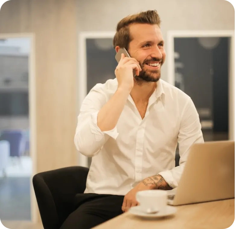 Smiling man in a white shirt talking on a smartphone while sitting at a desk with a laptop and coffee cup.