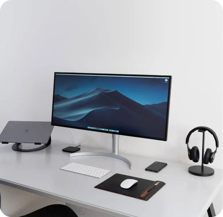 Minimalist workspace with curved monitor displaying a mountain screen, wireless keyboard and mouse on a black mat, smartphone, laptop on a stand, and headphones on a stand.