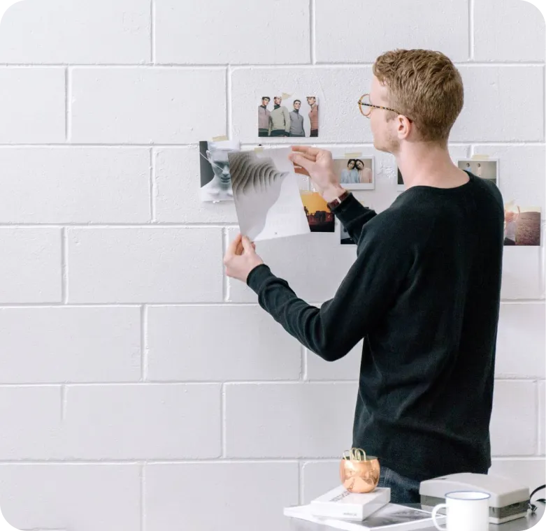 A man with glasses in a black sweater arranging photos and papers on a white brick wall.