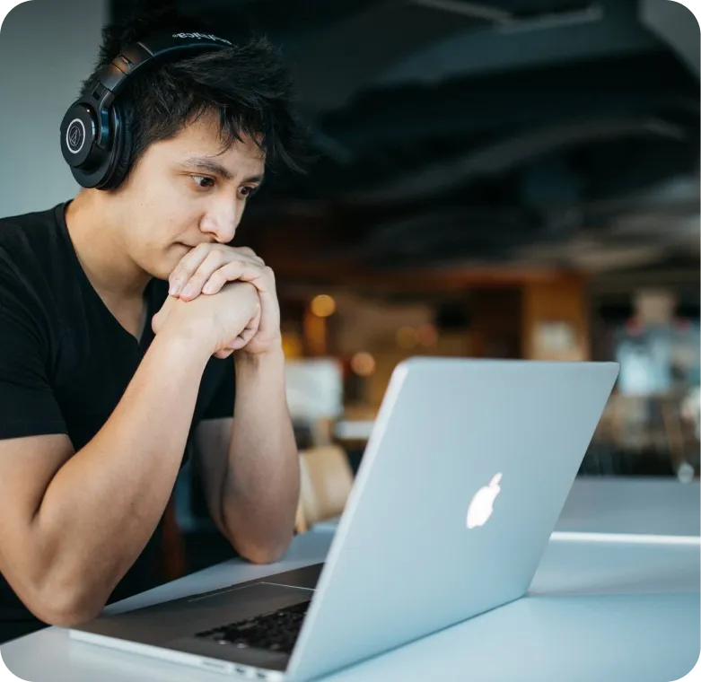 Young man wearing headphones looking thoughtfully at a laptop screen in a modern indoor setting.