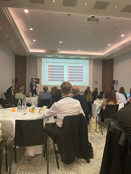 Three people sitting at a table, talking and laughing in a bright room with plants.