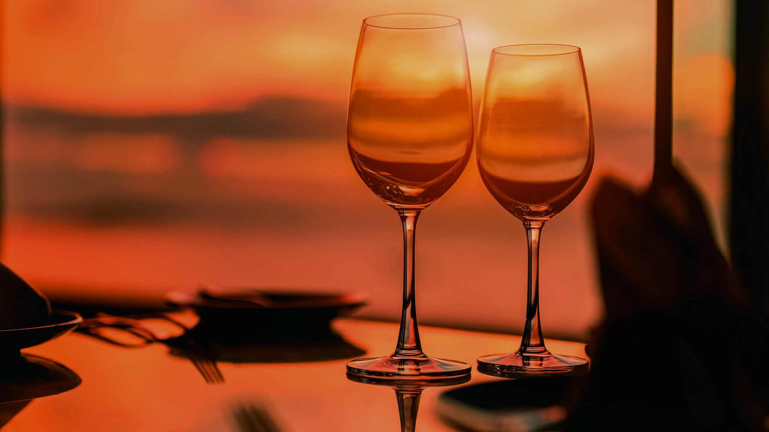 Two wine glasses catching a deep oxblood sunset over the sea at a candlelit table, quiet luxury dining