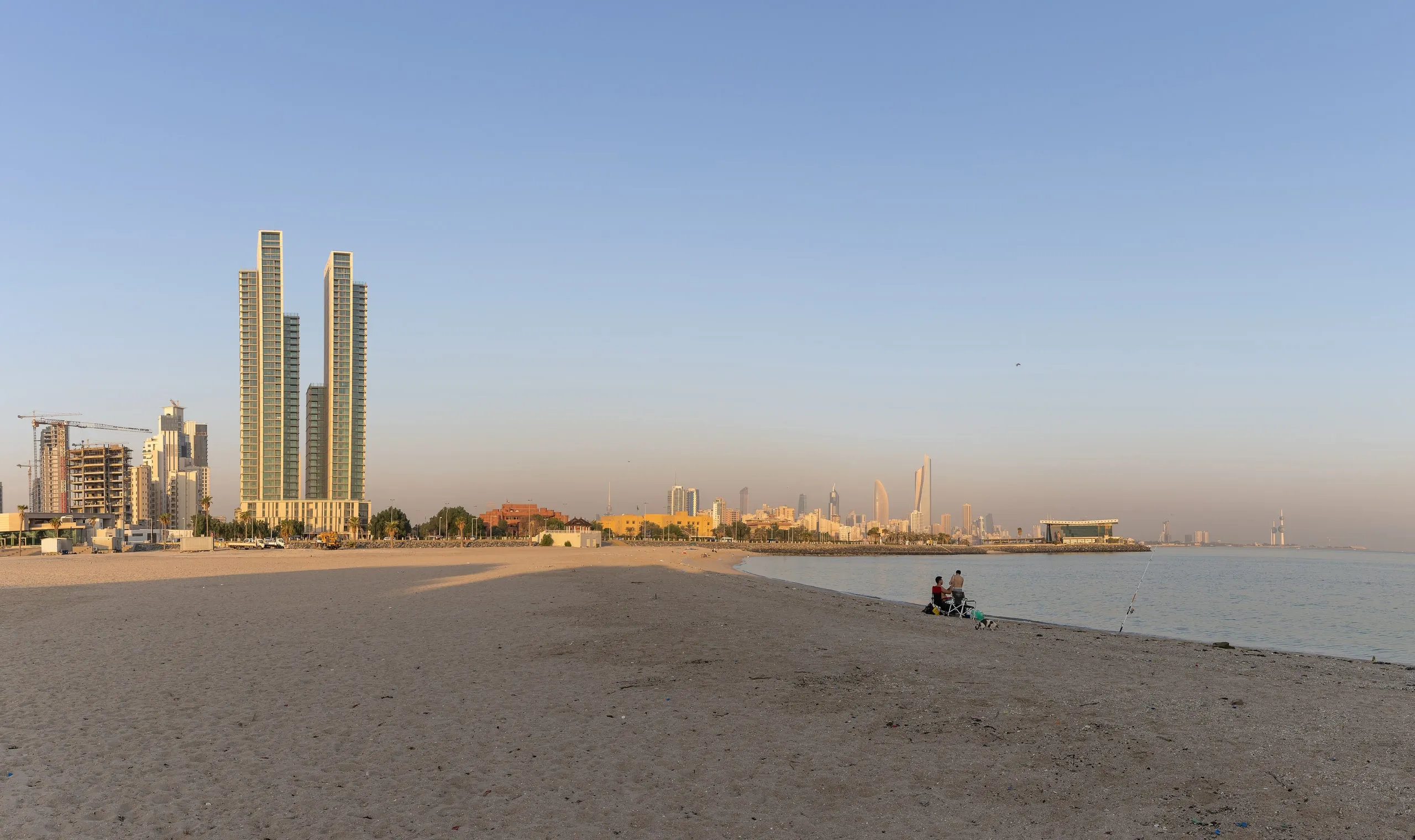 Twin high-rise towers of Hessah Al Mubarak in Kuwait City seen from the beach, with the coastal skyline stretching along the shoreline at dusk