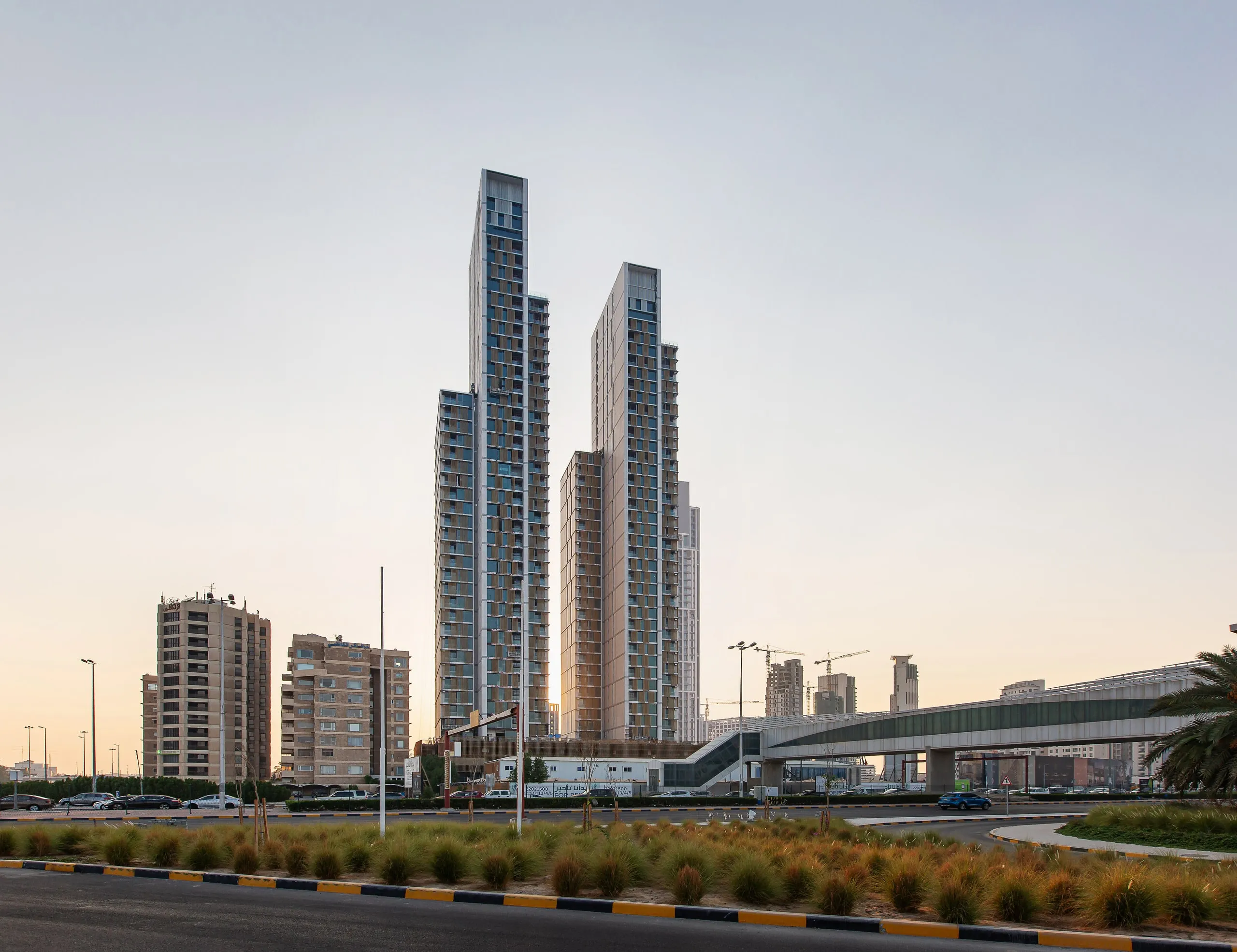 Twin high-rise towers of Hessah Al Mubarak in Kuwait City, tapering at their crowns above a podium, seen at dusk beyond a highway roundabout
