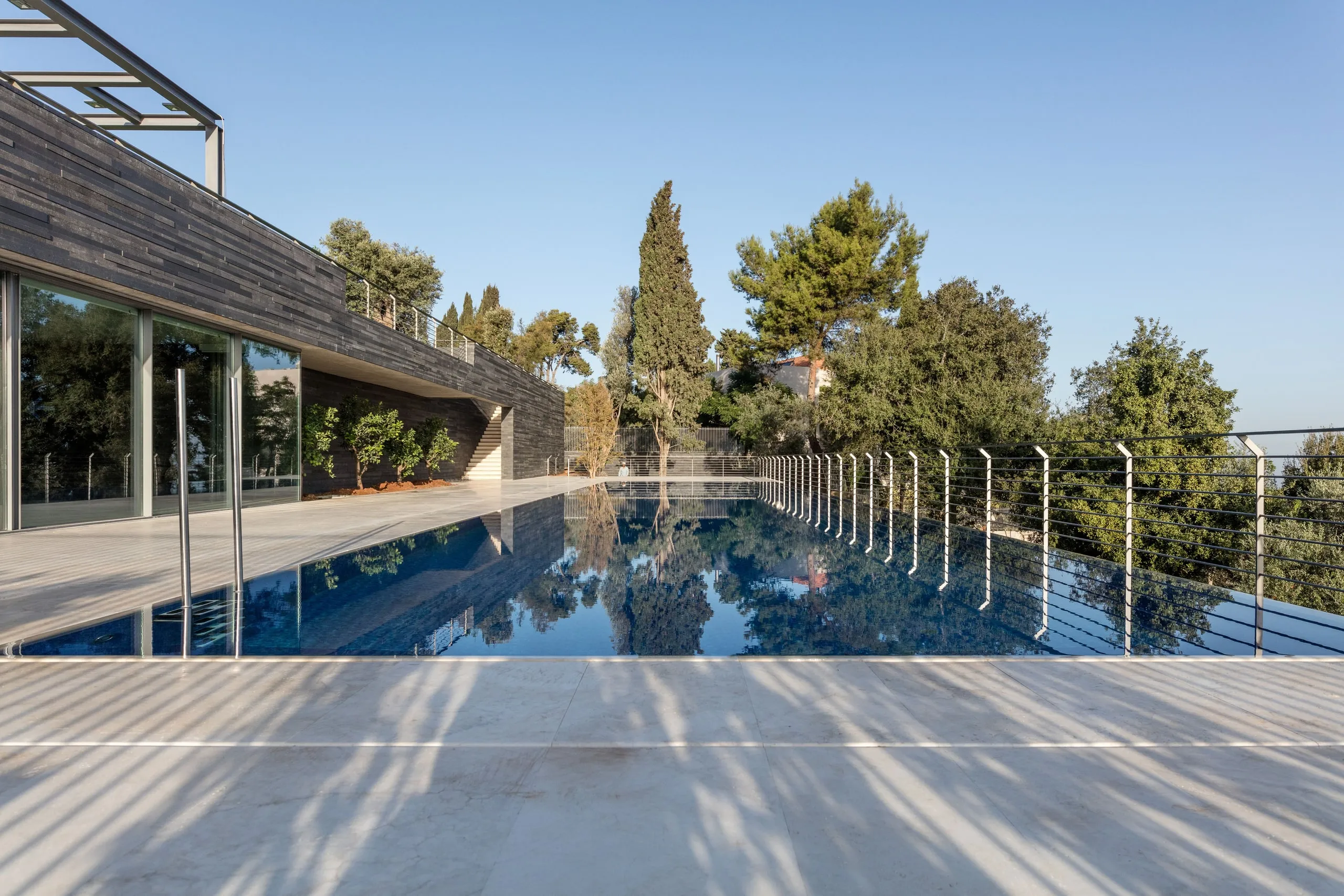 Long lap pool at AY House in Yarze, lined by a slatted dark timber facade and cable railing, with pine trees reflected in the still water