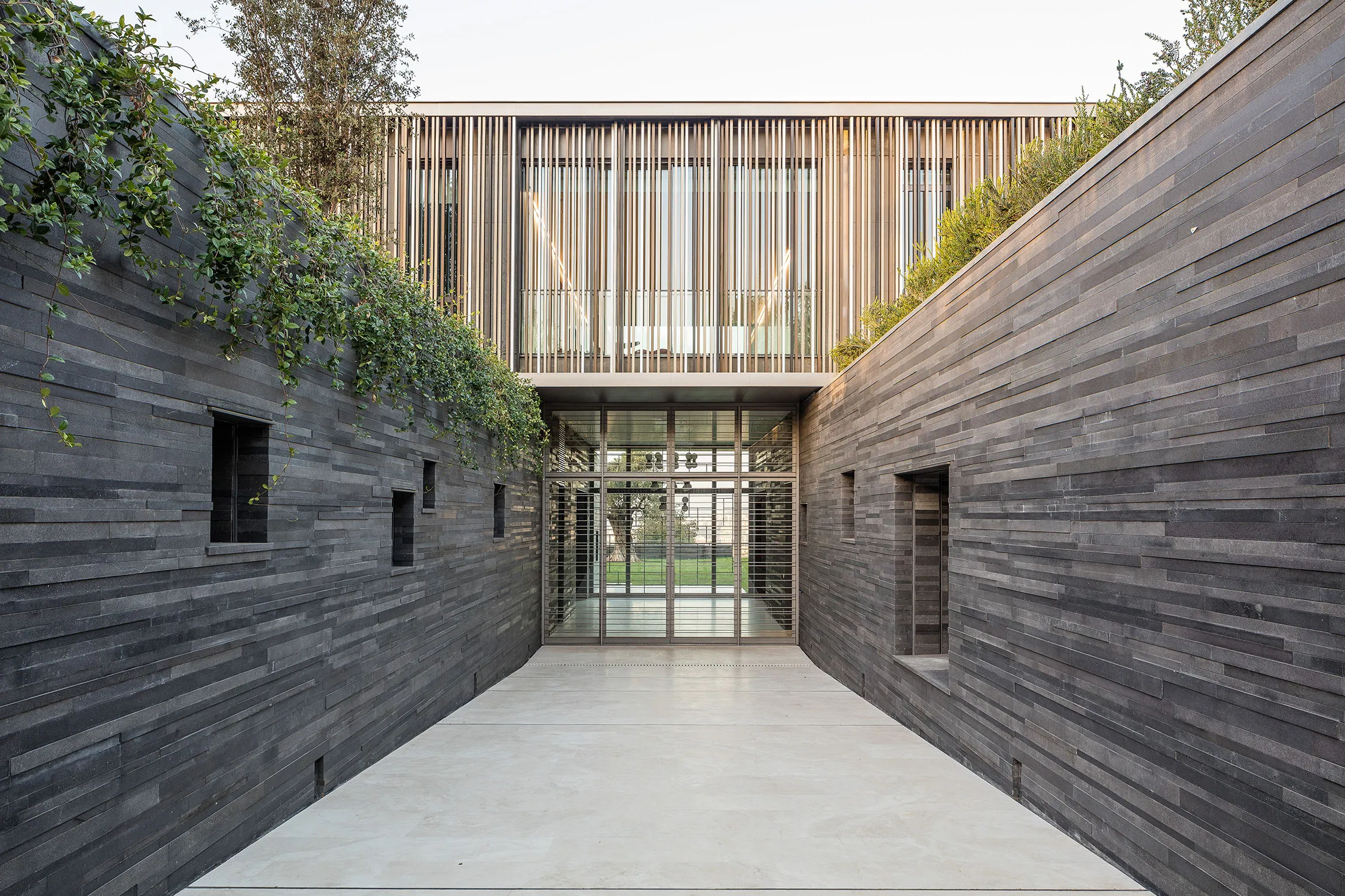 Basalt-clad entrance corridor of AY House in Yarze leading to a glazed doorway, with a timber-louvered upper floor cantilevering above