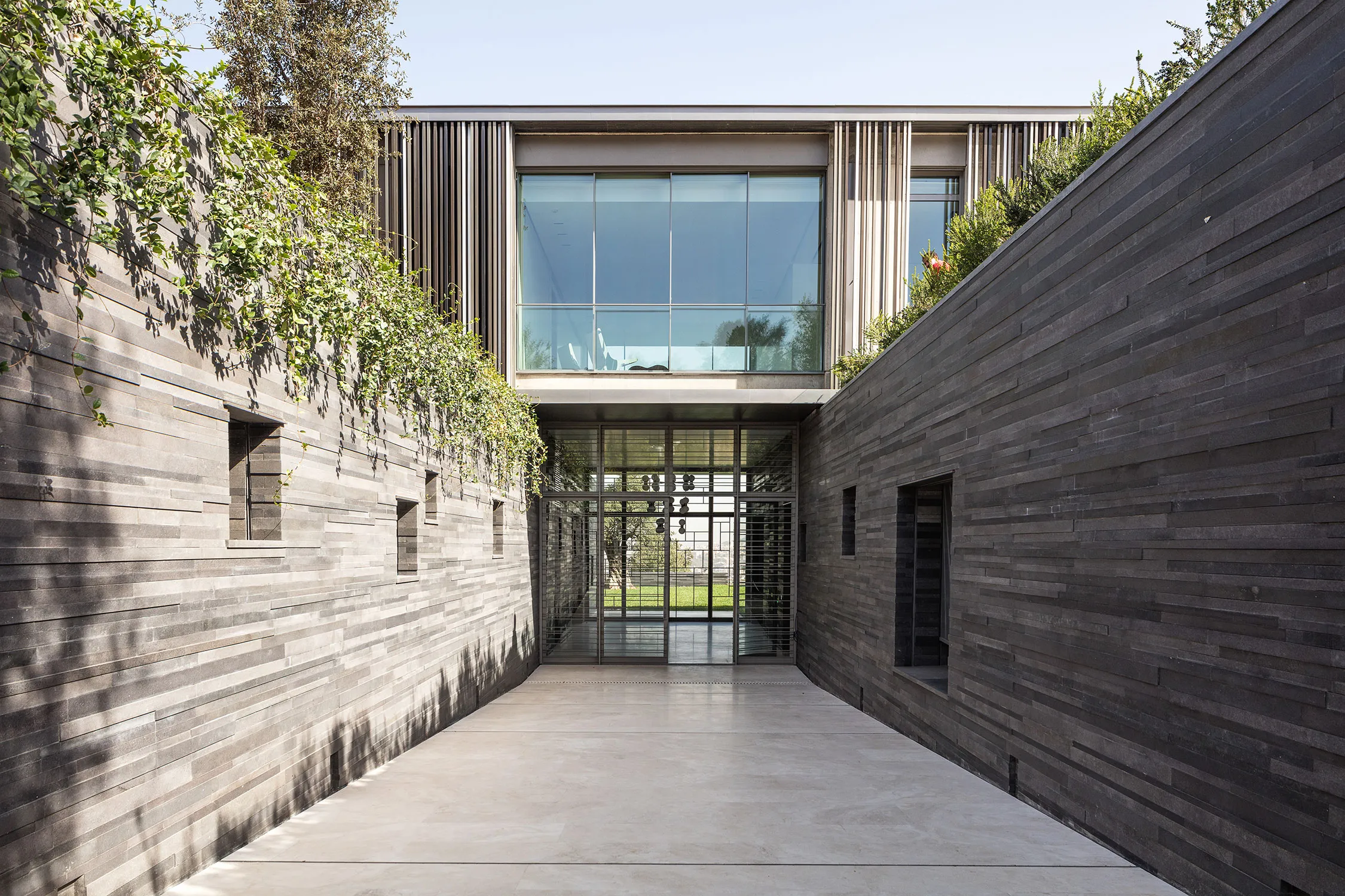 Entrance courtyard of AY House in Yarze, dark basalt-clad walls lined with climbing plants leading to a glazed doorway and garden beyond